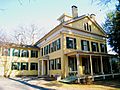 A yellow Federal style house with white trim and green shutters. The front of the house has a covered porch, but the main entrance is on the side of the house. It is protected by a small portico.