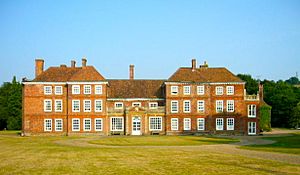 Large red brick building with sash windows of 15 bays, the 3 central bays only 2.5 stories, the outer six on each side of 3.5 and the final bay of each side filled in with brick. On a trimmed lawn