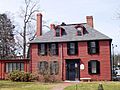 A 2+ story colonial house with two chimneys. It is painted red and has black shutters.