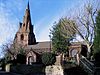 A church in red sandstone, the steeple with pinnacles to the left and the body of the church, with round-headed windows to the right. In front of the church are trees, a wall, and steps leading to the churchyard which contains a table tomb.