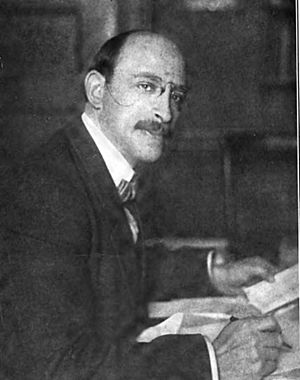 A 42-year-old man wearing a bowtie and suit leans over a desk as he writes