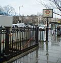 A white sign reading "REGENT'S PARK STATION BAKERLOO LINE" in black letters with people walking behind it all under a light blue sky