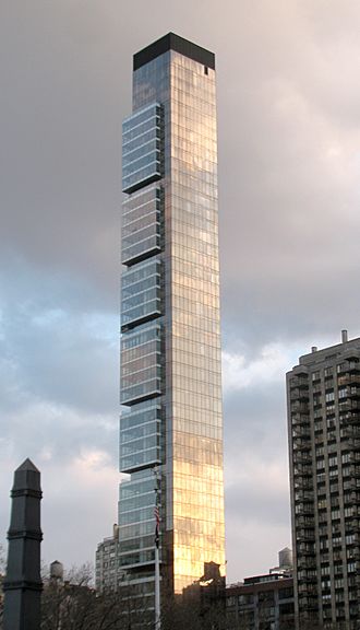 A tall, thin building with some slight squarish projections at its higher levels seen from between some trees.