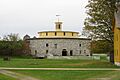 This color photo shows a round barn made of fieldstone. It is topped by a mustard yellow wood frame multi-sided section that is smaller, and then a small turret.