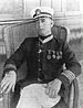 A white man sitting relaxed in a wicker chair. He is wearing a white cap, light–colored pants, and a dark jacket with four round medals on the left breast and a star–shaped medal around the neck.