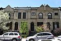 A cream-colored two-story smooth-surfaced building with pointed projections and parapets above its roofline, arched doorways and stoops. In front are small trees in various stages of spring blossoming, and two silver parked SUVs.