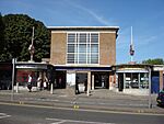 A brown-bricked building with a blue sign reading "EASTCOTE STATION" in white letters and people walking in front all under a blue sky with white clouds