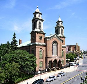 A brick building with two tall towers topped with copper, seen from its right, across a city street, at some height. Other buildings are visible on the right of the image.