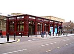 A red-bricked building with a series of white signs together reading "KILBURN PARK STATION" in red letters and people walking in front all under a white sky