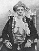 A black-and-white photograph of a man with a white beard wearing a turban, a dark jacket, a white shirt, and a belt and sitting on a chair