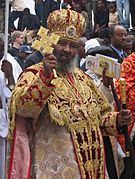 Abune Paulos dressed in religious ceremonial garb including mitre that is red with an abundance of gold. He is holding a staff and golden processional cross.