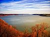 View of a very wide river lined with bare trees in reddish light under a blue sky streaked with clouds