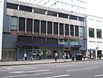 A grey building with blue panels and a dark blue, rectangular sign reading "LANCASTER GATE STATION" in white letters and people in front