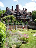 A wooden-shingled house with many gables has a formal garden in front of it.