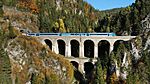 A photochorm picture of the a curving railroad bridge coming out of a tunnel bored into a large mountain.  In the background another bridge and mountains are visible.