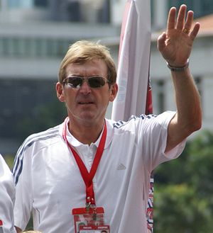 Colour photograph of Dalglish in Singapore, 2009. He is dressed casually, wearing sunglasses and has his hand raised