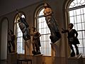 An interior view of figures that once graced the bows of wooden sailing ships. High windows flank two female figures that are mounted on the wall, and three male figures stand on pedestals in front of the windows.