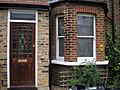 A brown brick building. Visible is a door at left, with a bowfront window at right.