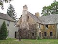 The rear of a 2+ story stone house has a wooden addition extending off to the left. An old-fashioned water pump with bucket is near the door.