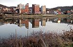 Modest skyscrapers, their images reflected in the water, line the bank of a wide placid river.