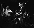 The band Flipper is performing at a club. From left to right are the singer, drummer and electric guitarist. The singer is seated on a stool, and he is holding a pair of crutches.