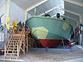 An open boat painted with camouflage colors sits inside a museum building. Wooden steps lead up to a platform from which the interior of the boat can be viewed. Flags hang from poles mounted on the left well.