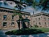 A flagpole and pine tree in front of a large two story stone building with many windows