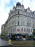 A white building with a rectangular, dark blue sign reading "KNIGHTSBRIDGE STATION" in white letters all under a light blue sky with white clouds