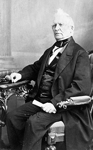 Phot of fair-skinned man with white hair, tuft in front, wearing mid-Victorian business suit, sitting at a table