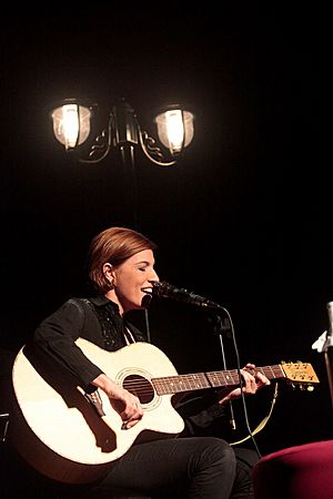 Lúcia Moniz seated on a darkened stage, playing acoustic guitar and singing into a microphone