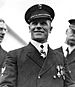 Head and shoulders of a smiling white man wearing a dark peaked cap with the letters "U.S.N" on the front and a dark jacket with three medals on the left breast and a star-shaped medal hanging from a ribbon around his neck.