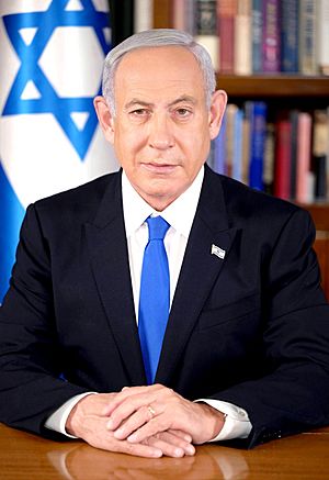 Portrait of Benjamin Netanyahu, a middle-aged man, wearing a suit with a blue tie, sitting in front of an Israeli flag.