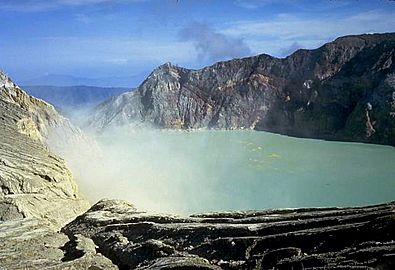 A photograph depicting a blue sky with white clouds at the top, a light grey lake in the middle, and dark grey rocks surrounding the lake.