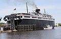 A large black ship with white upper decks rests at a berth, its rear surrounded by wooden posts.  The rear access to its vehicle decks is opened upward.