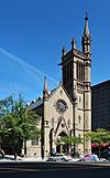 A Gothic stone church with a pointed facade and a tower on the right, with a smaller tower rising from its own right, seen from across a city street