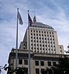 Ground-level view of an Art Deco-style skyscraper with a tan facade; the building is adorned with a thick, gray spire. In the foreground, two flags are visible.