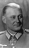 The head of a man, shown in semi-profile. He wears a military uniform with a military decoration in shape of an iron cross displayed at the front of his shirt collar. His hair is combed to back.