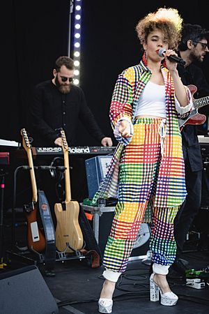 Pale skinned black female wearing platform shoes and a multi coloured patchwork trouser suit singing on stage with a background of racked spare guitars, one guitarist and a keyboard player