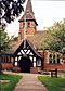 A path leads to a black-and-white timber-framed porch with a flagpole. The body of the church extends on both sides and beyond the porch is a broach spire with a clock face