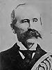 Head of a white man with a large mustache and receding hairline wearing a dark suit, white shirt, and dark tie. Over the bottom right of the portrait is a decorative design.