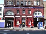 A red-bricked building with a blue sign reading "GOODGE STREET STATION" in white letters and several people walking in front all under a blue sky