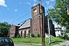 A brown brick building with gothic detailing, buttresses and a square tower on the front right seen from the middle of a nearby intersection.