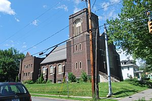 A brown brick building with gothic detailing, buttresses and a square tower on the front right seen from the middle of a nearby intersection.