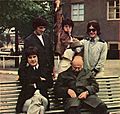 On a bench in a park sit five men, two seated and three standing behind the bench. Clockwise from left is a man in a black suit with Khaki pants; a man in a black suit with black pants; a man wearing a brown coat with Khaki pants and raising his arm jokingly as if to stab the sleeping man below him; a man, wearing a grey/light green coat and Khaki pants and holding out a hat above the sleeping man's head. The sleeping man is wearing green.