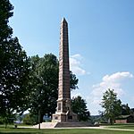 A stone obelisk monument against a blue sky and trees, with the statue of a pioneer in the front.