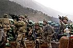 Seven soldiers in beige tactical gear huddle behind a row of green sandbags on a mountainside, pointing rifles in various directions