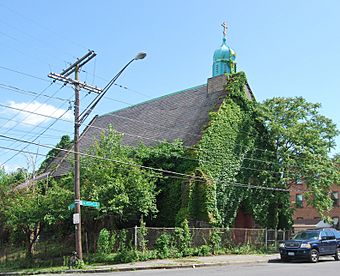 An ivy-covered building with a pointed roof and a small green dome with a cross seen from across a street, with a car parked in front. There is a chainlink fence around it and the ground slopes downwards towards its rear. A street sign at the corner reads "Colonie" and "North Pearl".