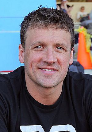 Photo is a profile head shot of Ryan Lochte, a white man with sandy brown hair and blue eyes, standing behind a microphone