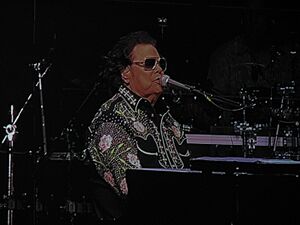 Ronnie Milsap in a sequined black suit and sunglasses, seated at a piano while singing into a microphone