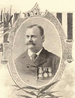 Head and shoulders of a white man with a mustache and receding hairline, wearing a dark suit and tie with three medals hanging from ribbons on the left breast. The portrait is surrounded by a decorative oval frame.